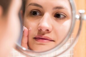A young girl in front of a bathroom mirror putting a spot treatment cream on a red pimple