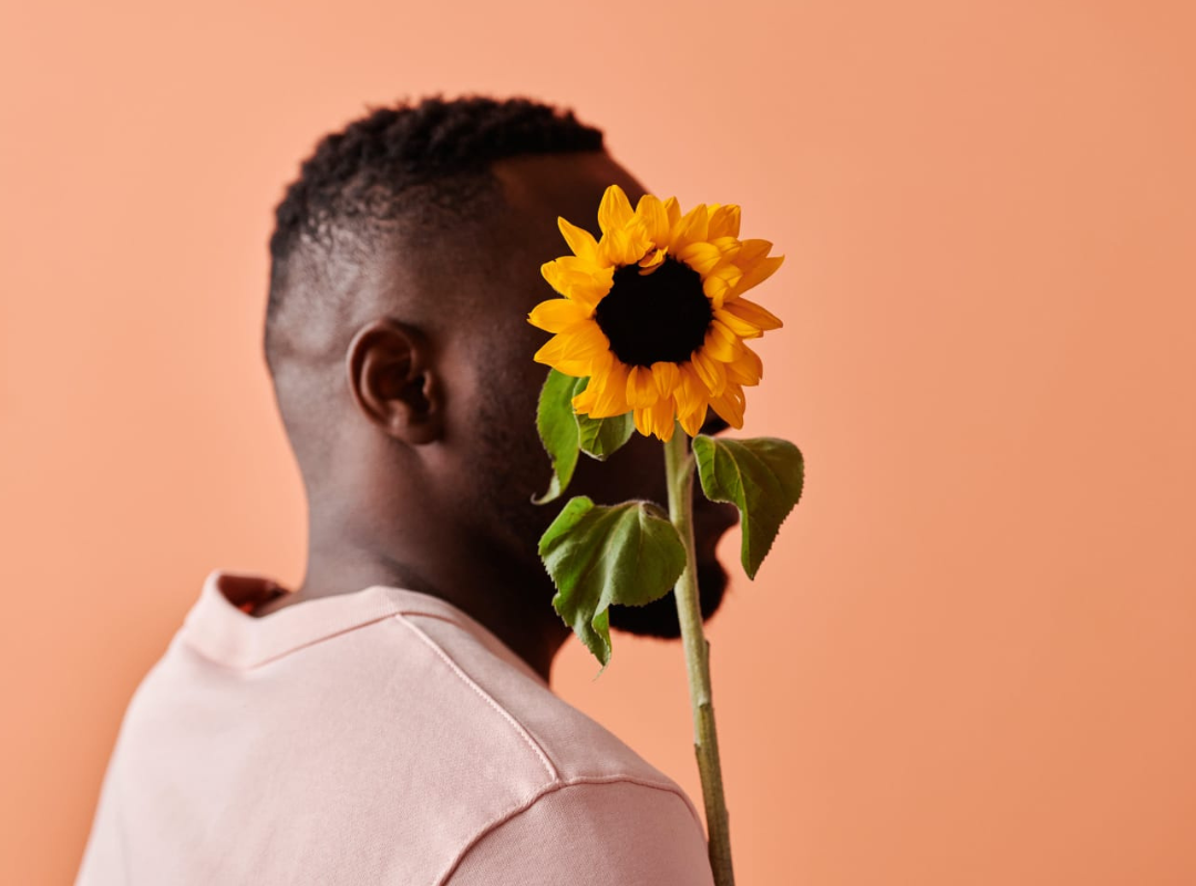 A fashionable person holding a sunflower against a peach background.