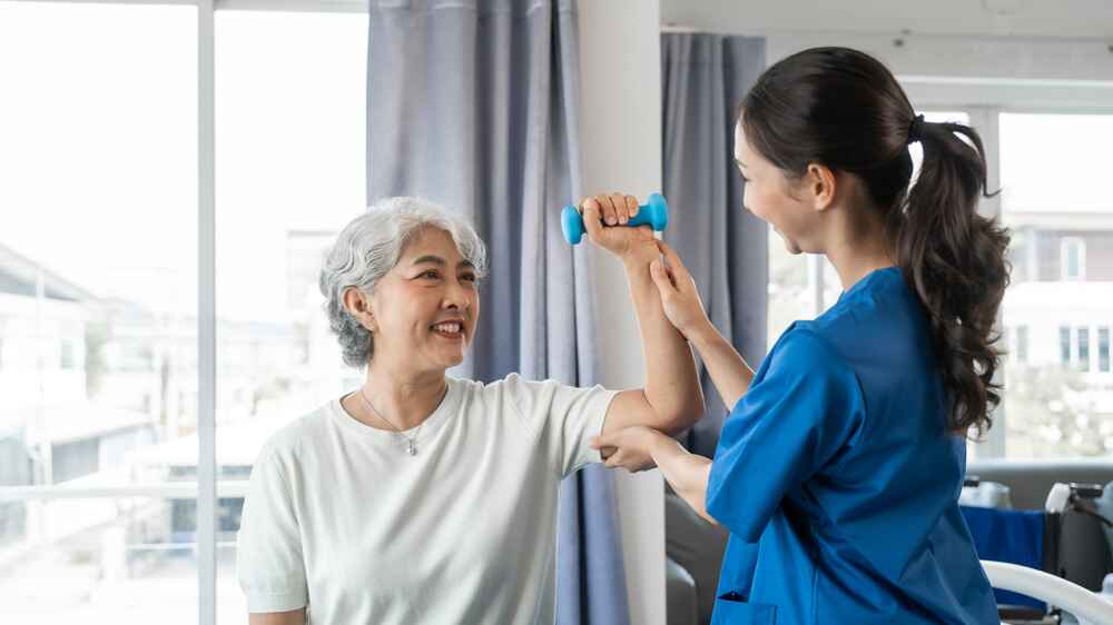 Young physiotherapist helping senior mature asian woman grey hair work out with dumbbells, to recover from injury at health centre in physical therapy session.
