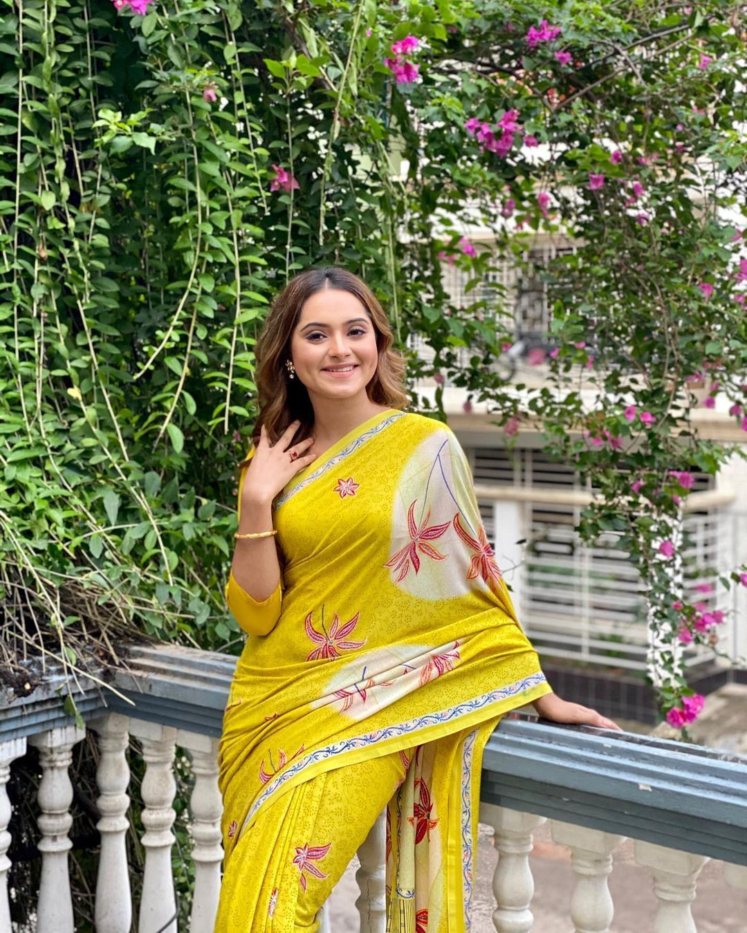 This may contain_ a woman in a yellow sari posing on a balcony with pink flowers behind her
