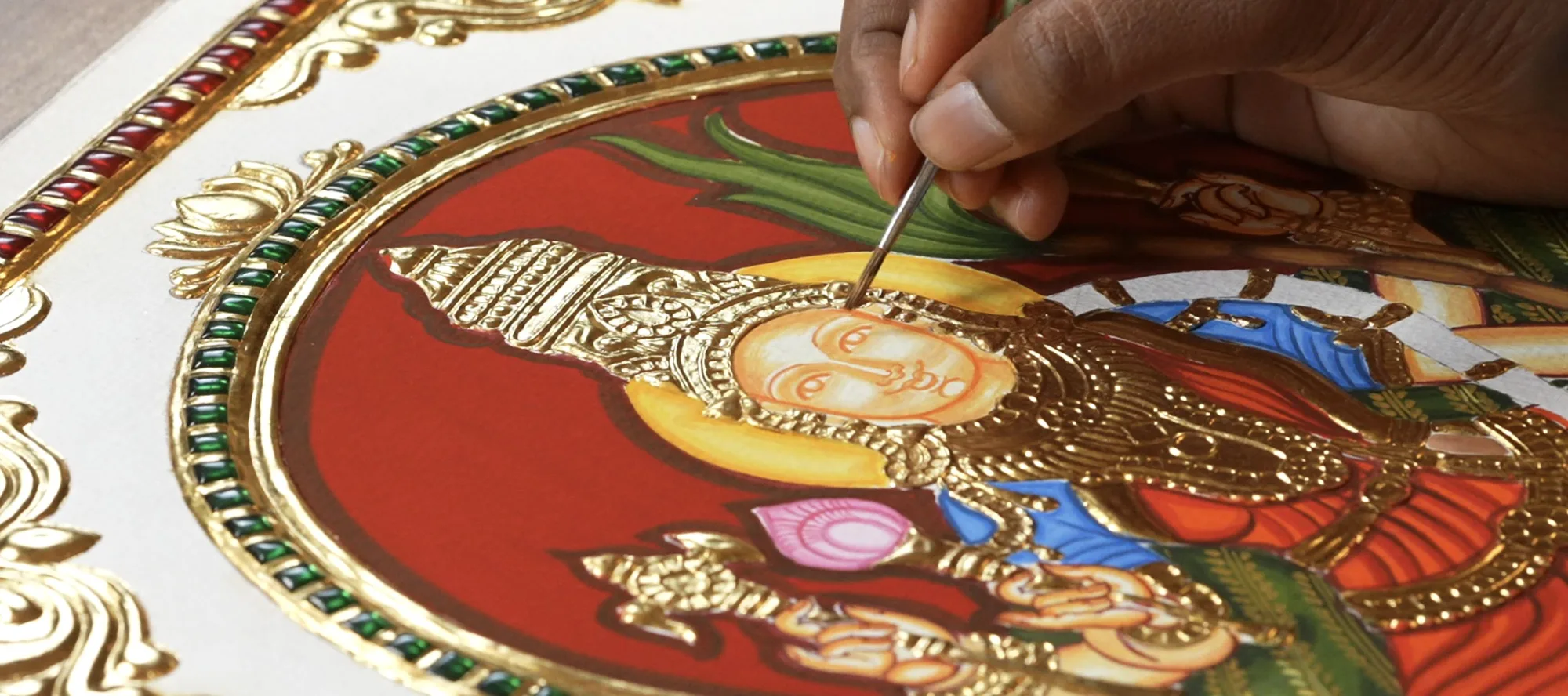 A fine brush adding detail to a Tanjore painting beside raised gold work