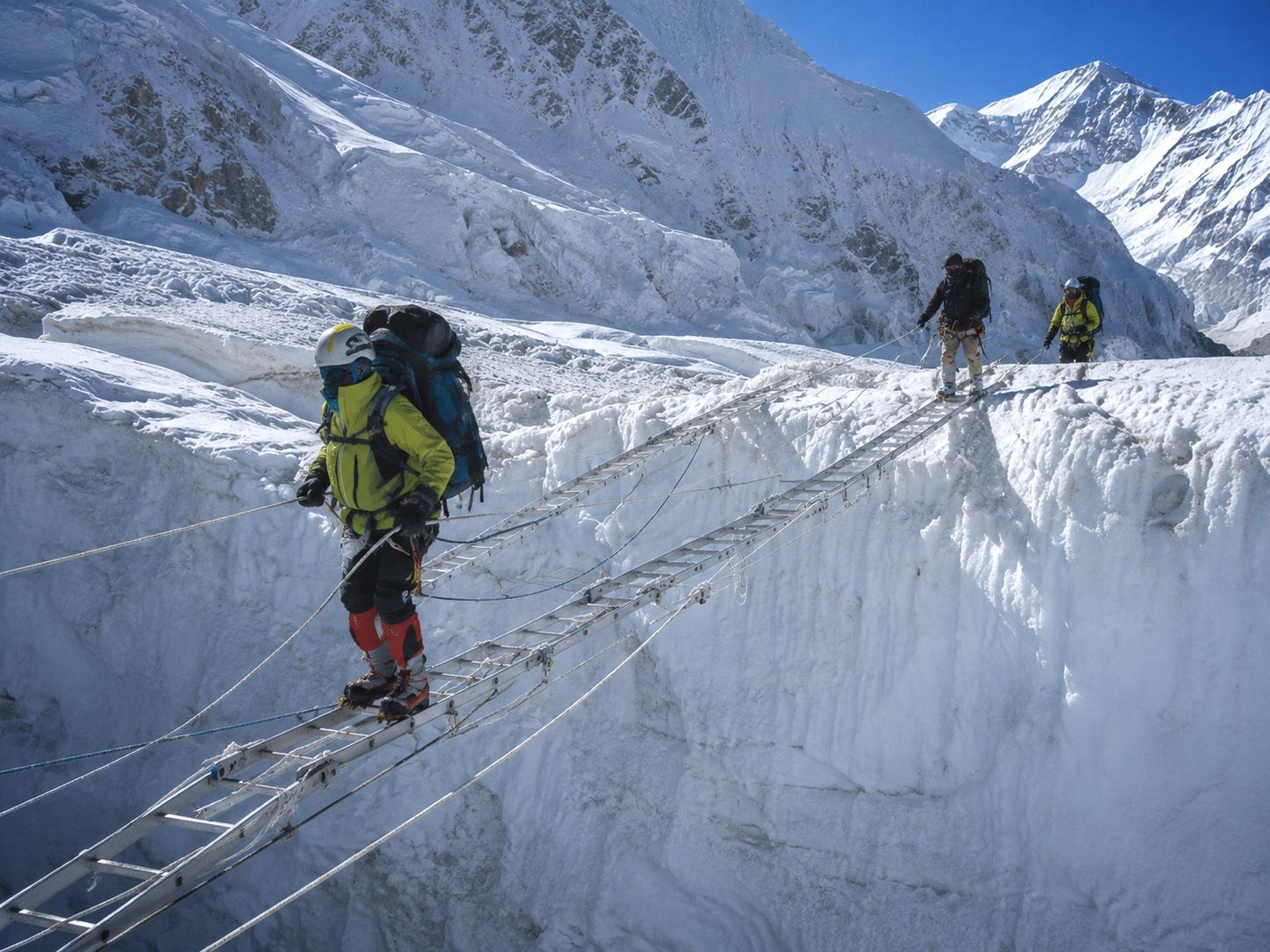 Mountaineers wearing helmets, crampons and heavy backpacks carefully cross a metal ladder spanning a deep ice crevasse in the Khumbu Icefall on Mount Everest as safety ropes are attached to the ladder, towering walls of snow and ice rise around them beneath a clear blue sky
