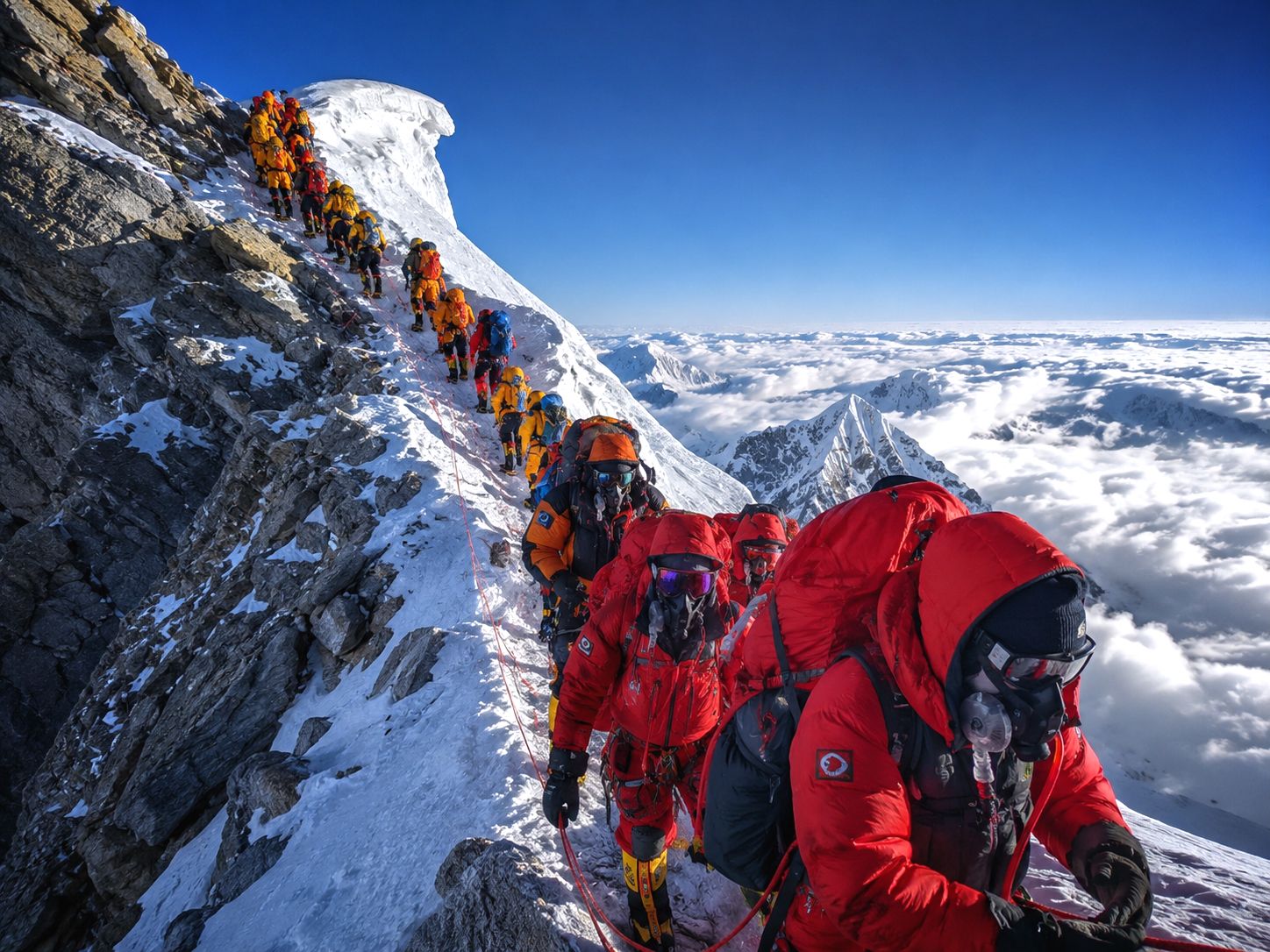 A long line of mountaineers in brightly coloured down suits and oxygen masks moves along a narrow snow-covered ridge high on Mount Everest; climbers are clipped to fixed ropes as steep rocky slopes drop away on one side, and cloud-covered Himalayan peaks stretch into the distance under a clear blue sky
