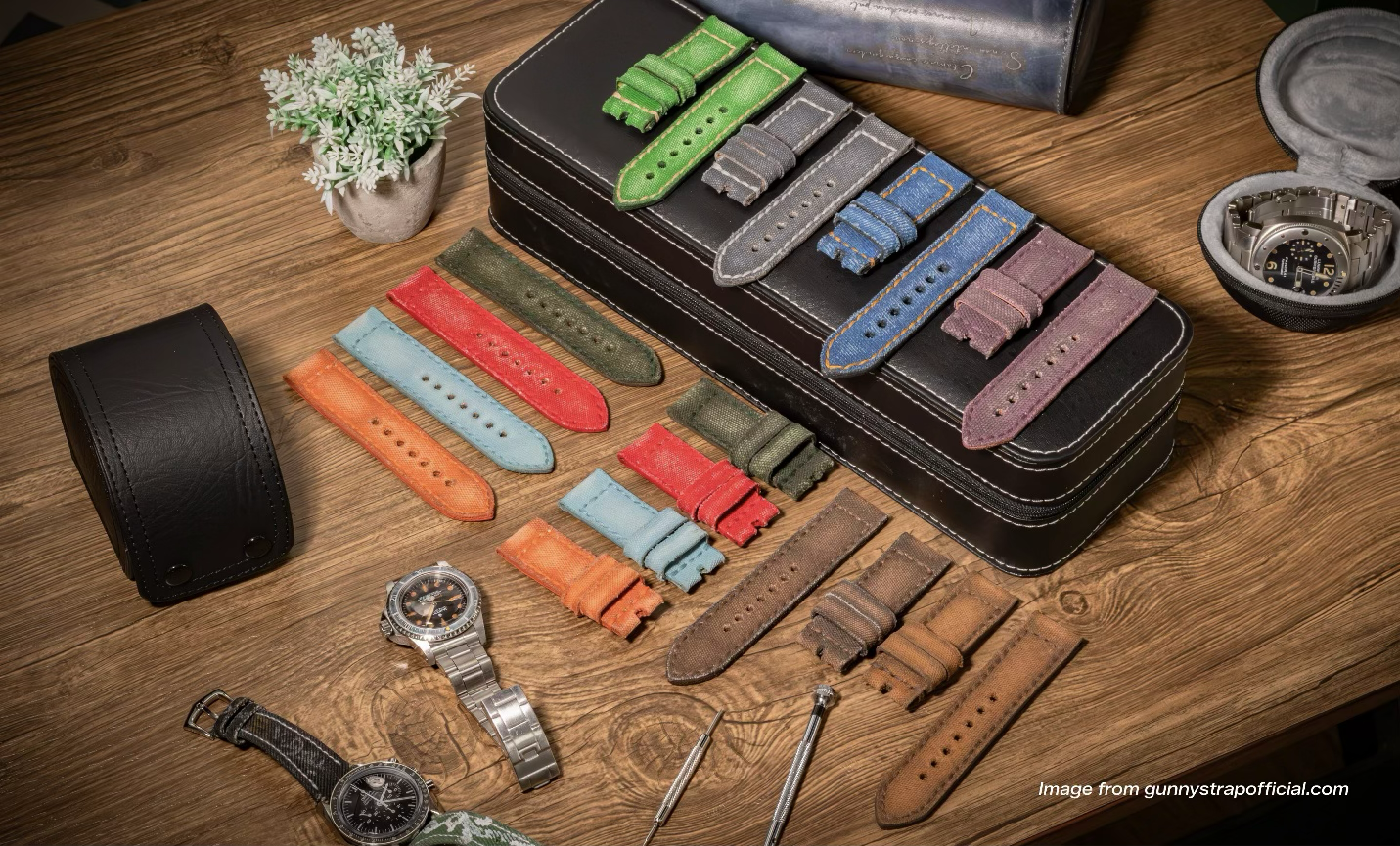 Koleksi strap kanvas Gunny Straps berwarna hijau, abu-abu, biru, ungu, merah, oranye, dan cokelat di atas meja kayu bersama beberapa jam tangan