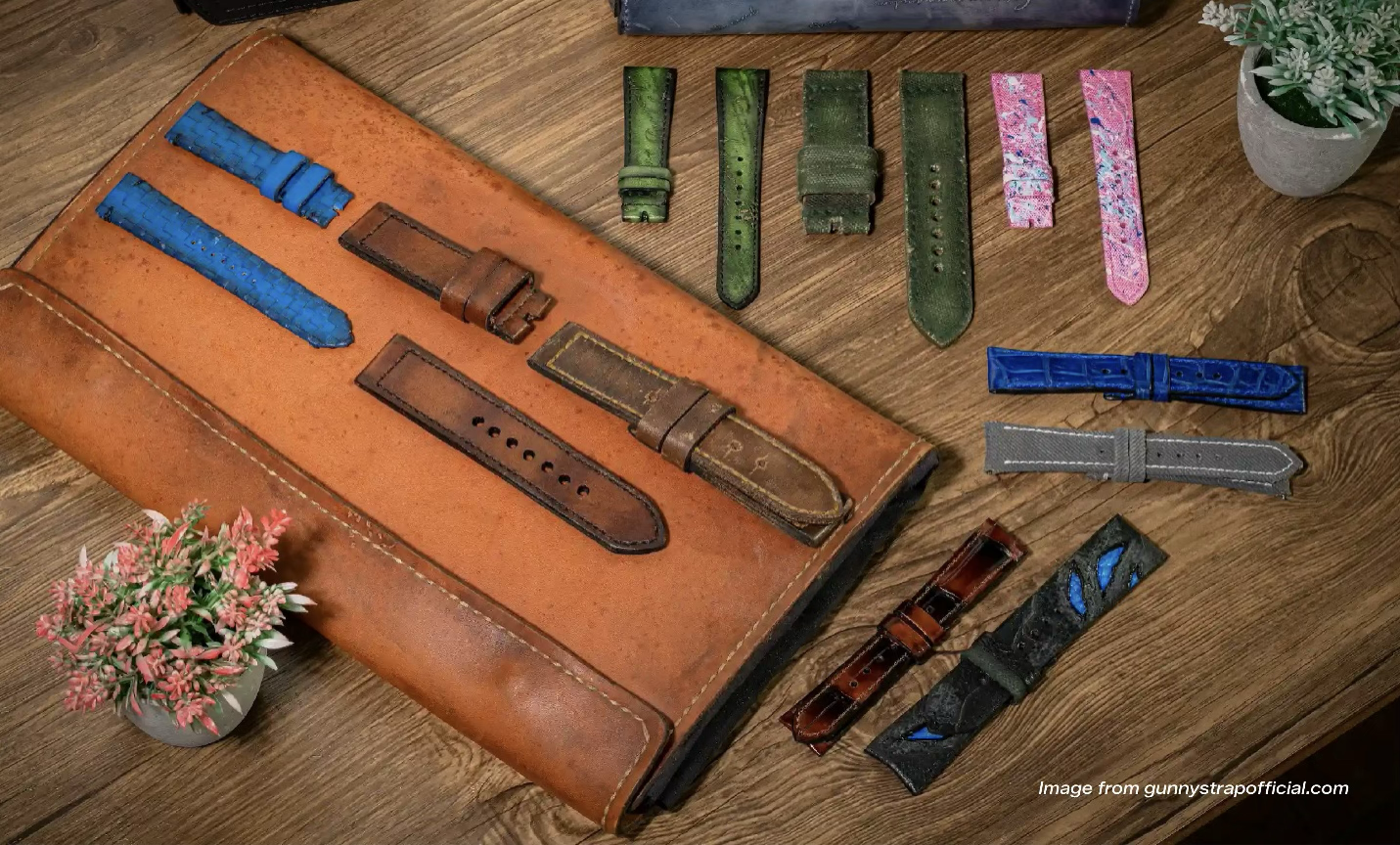 Berbagai strap Gunny Straps dari kulit, kanvas, dan kulit buaya dalam warna biru, cokelat, hijau, dan merah muda di atas tempat strap kulit dan meja kayu