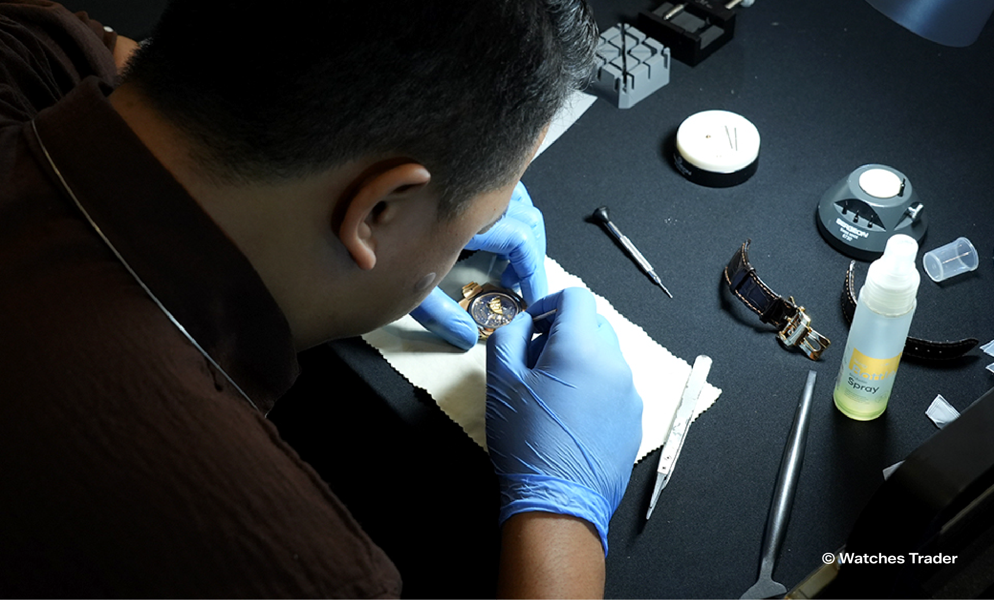 Teknisi Watches Trader bersarung tangan biru memeriksa jam tangan di atas kain putih, dengan obeng, pinset, dan peralatan lain di meja kerja