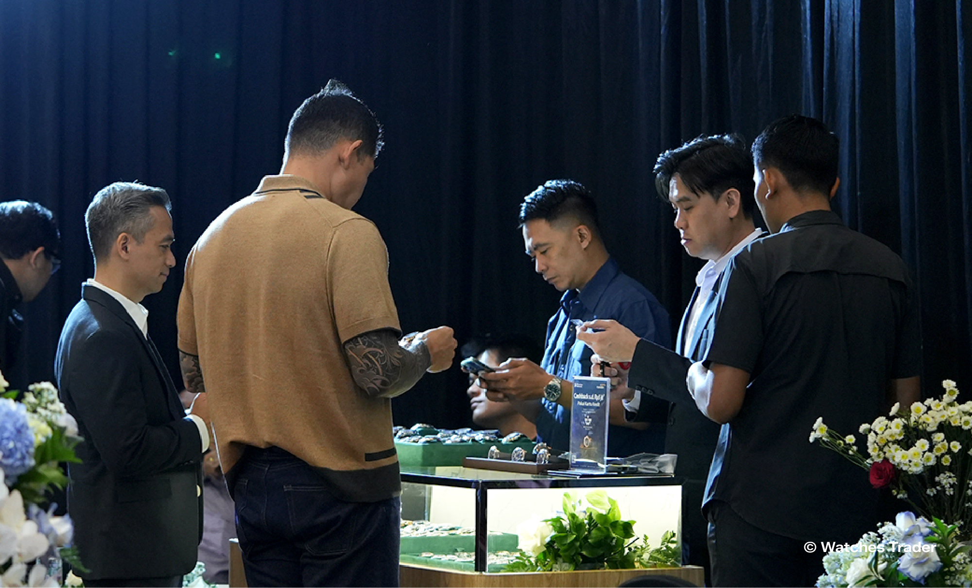 Sejumlah pengunjung dan staf Watches Trader berkumpul di sekitar etalase jam tangan yang dihiasi bunga, sambil berdiskusi dan melihat koleksi