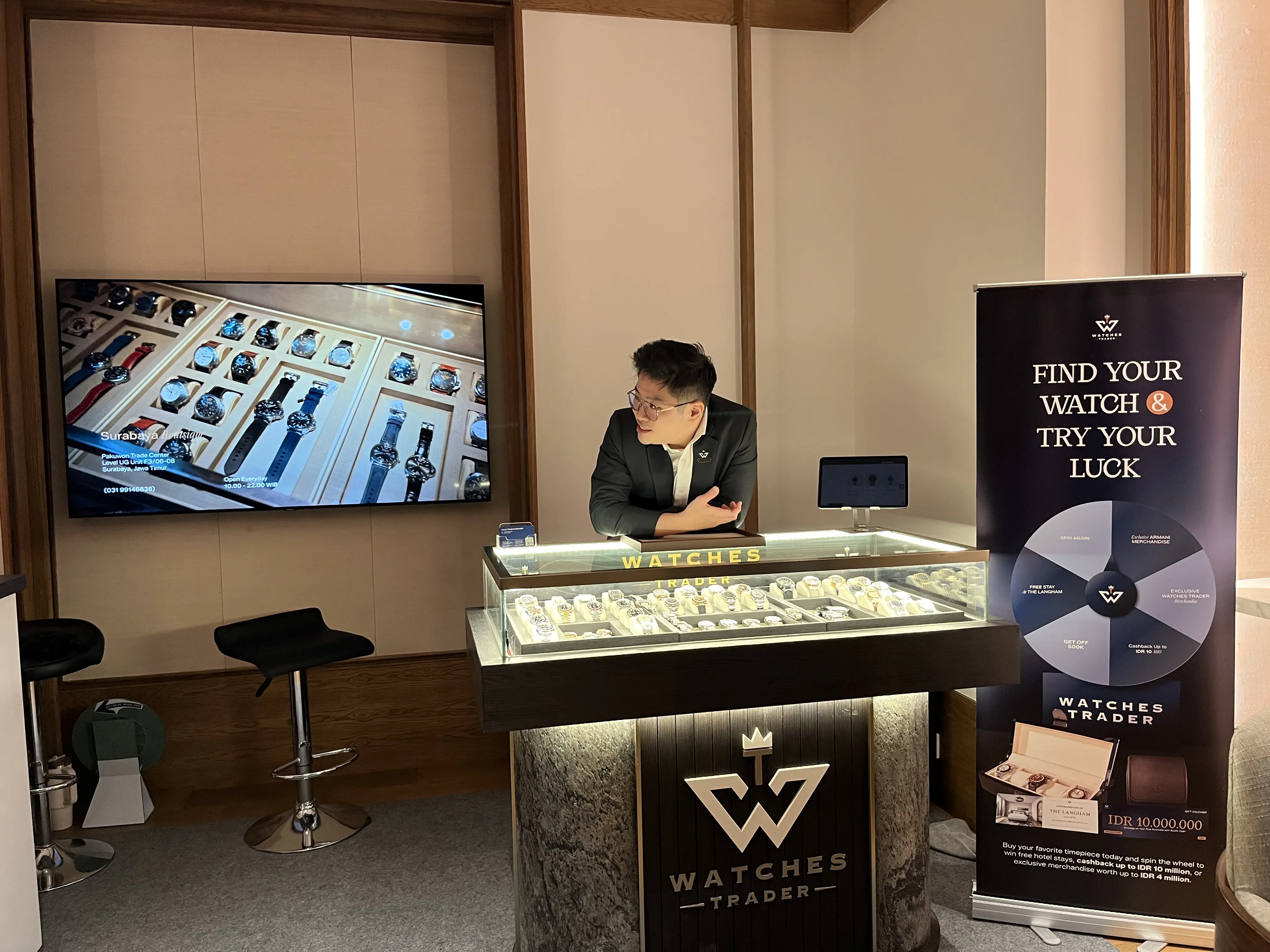 Staf Watches Trader di balik etalase jam tangan, dengan banner “Find Your Watch & Try Your Luck” berisi roda hadiah Spin the Wheel di sampingnya