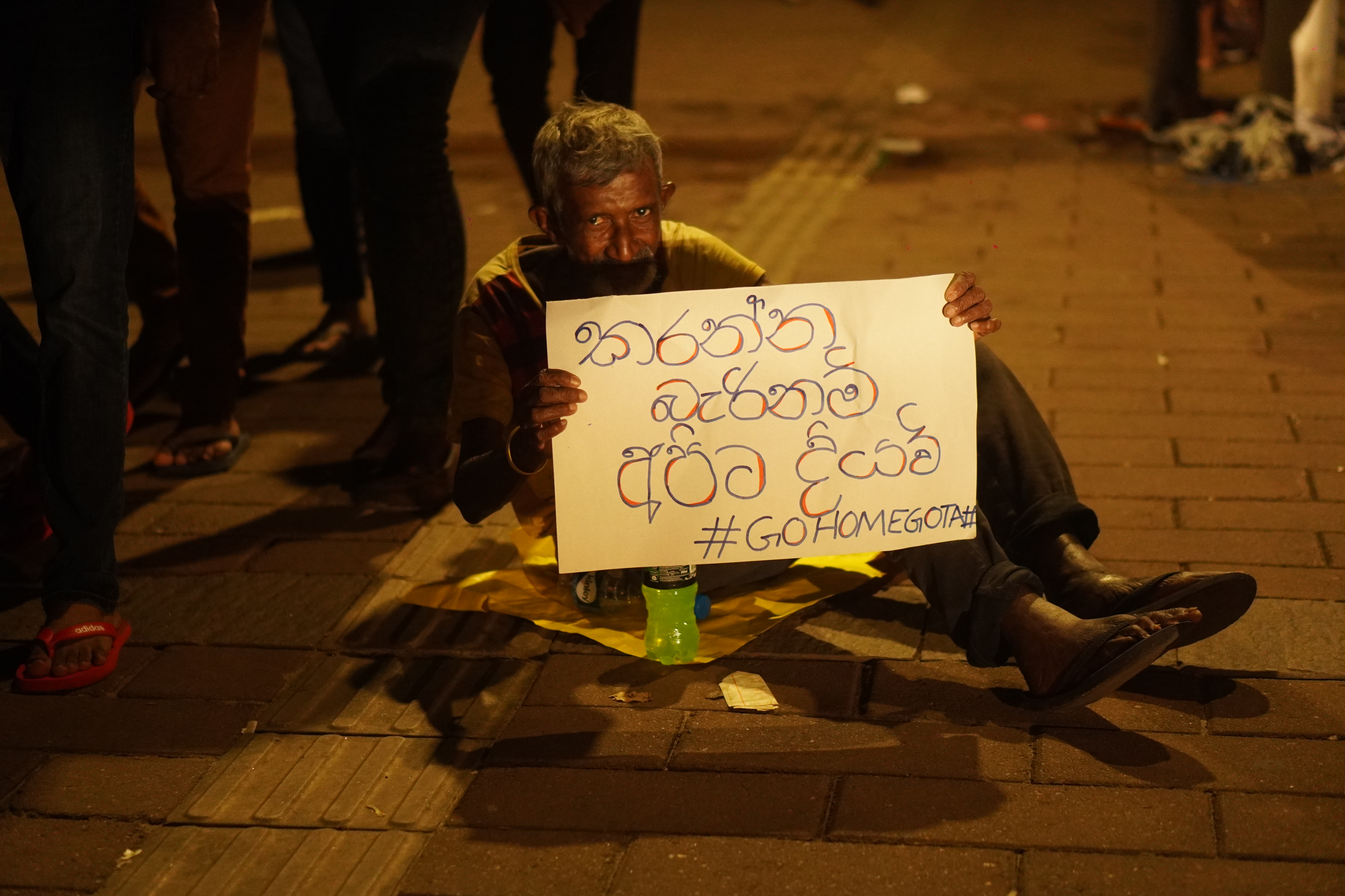 An elderly man seated on the ground holds a sign that says “If you can’t, then give it to us #GoHomeGota,” at a protest against the Rajapaksa administration in Galle Face, 20 April 2022. Photo by Riyal Riffai.