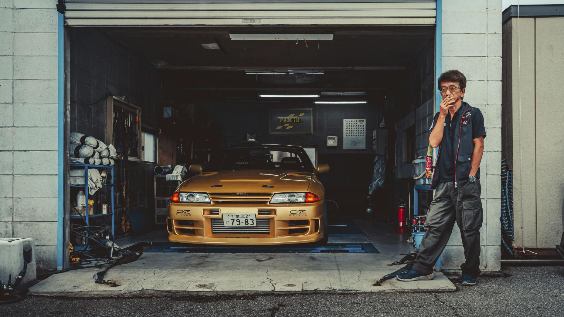 Smoky Nagata, founder of Top Secret, standing beside a gold Nissan Skyline GT-R R32 in a garage, symbolizing JDM tuning culture.