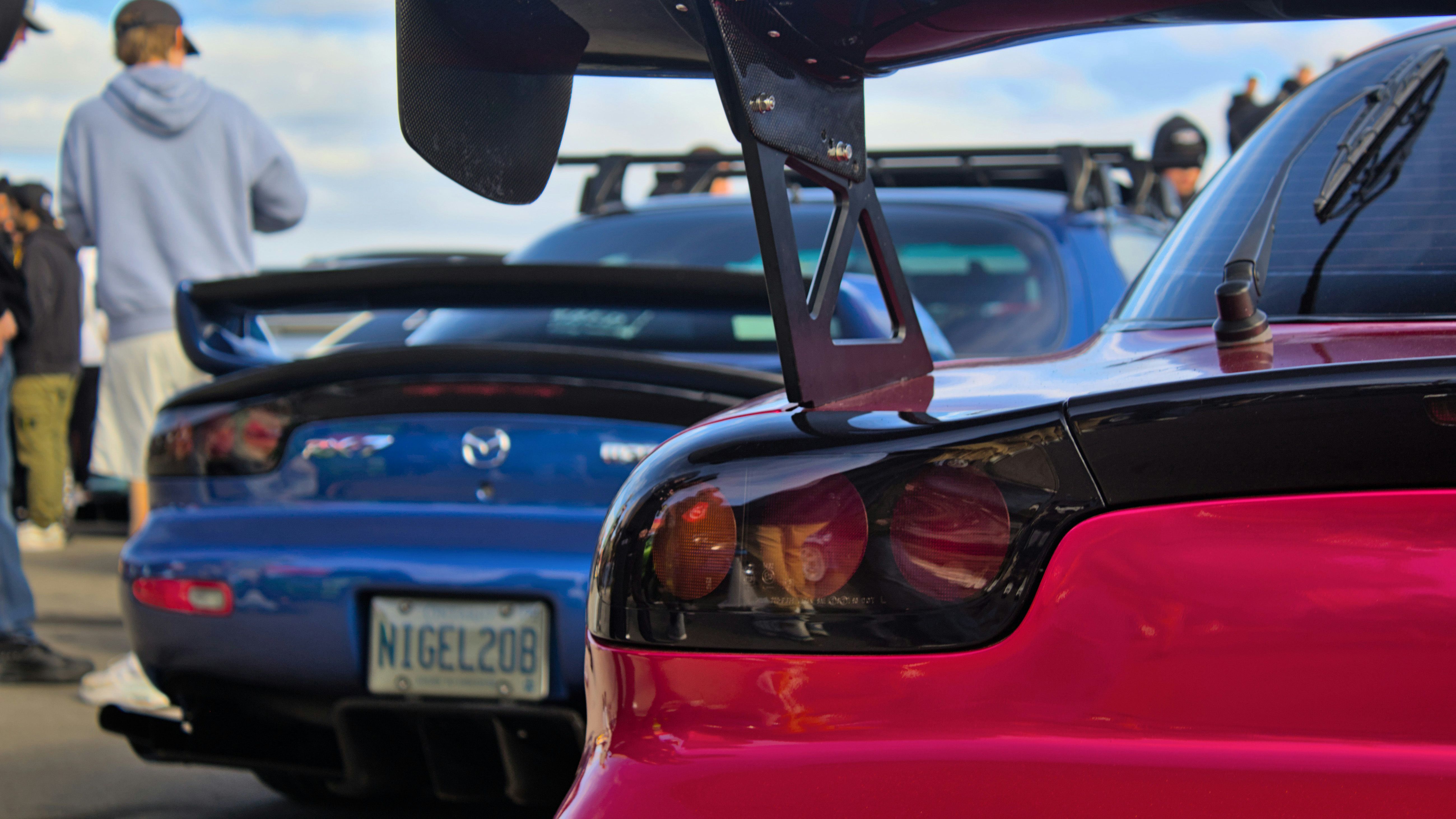 Rear view of two Mazda RX-7s, featuring aggressive aftermarket wings and iconic round taillights. A perfect scene for JDM enthusiasts, capturing the essence of rotary power and street culture.