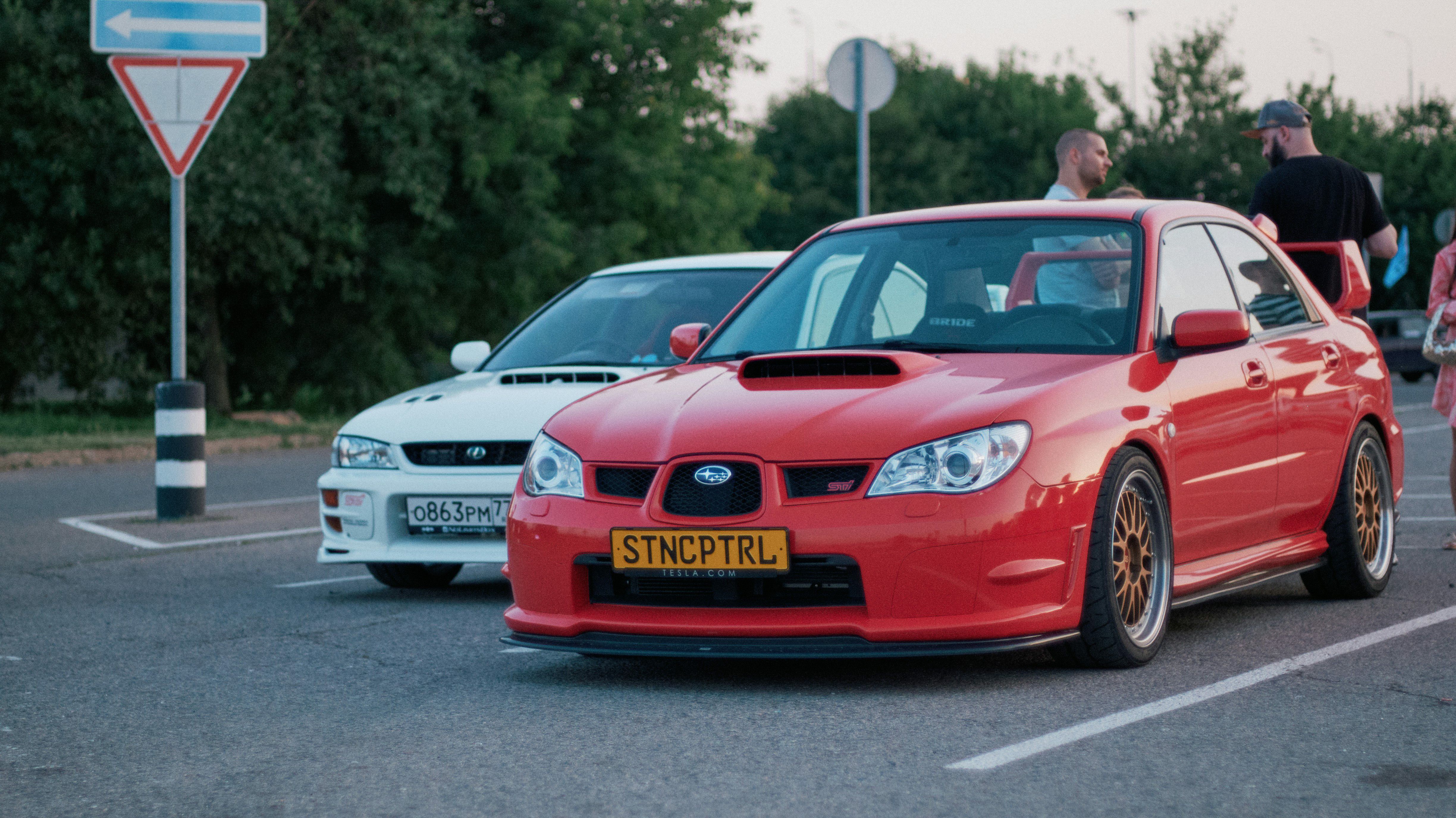 Red Subaru Impreza STI with gold wheels and aggressive stance, parked beside a white JDM car, showcasing iconic Japanese performance at a car meet.