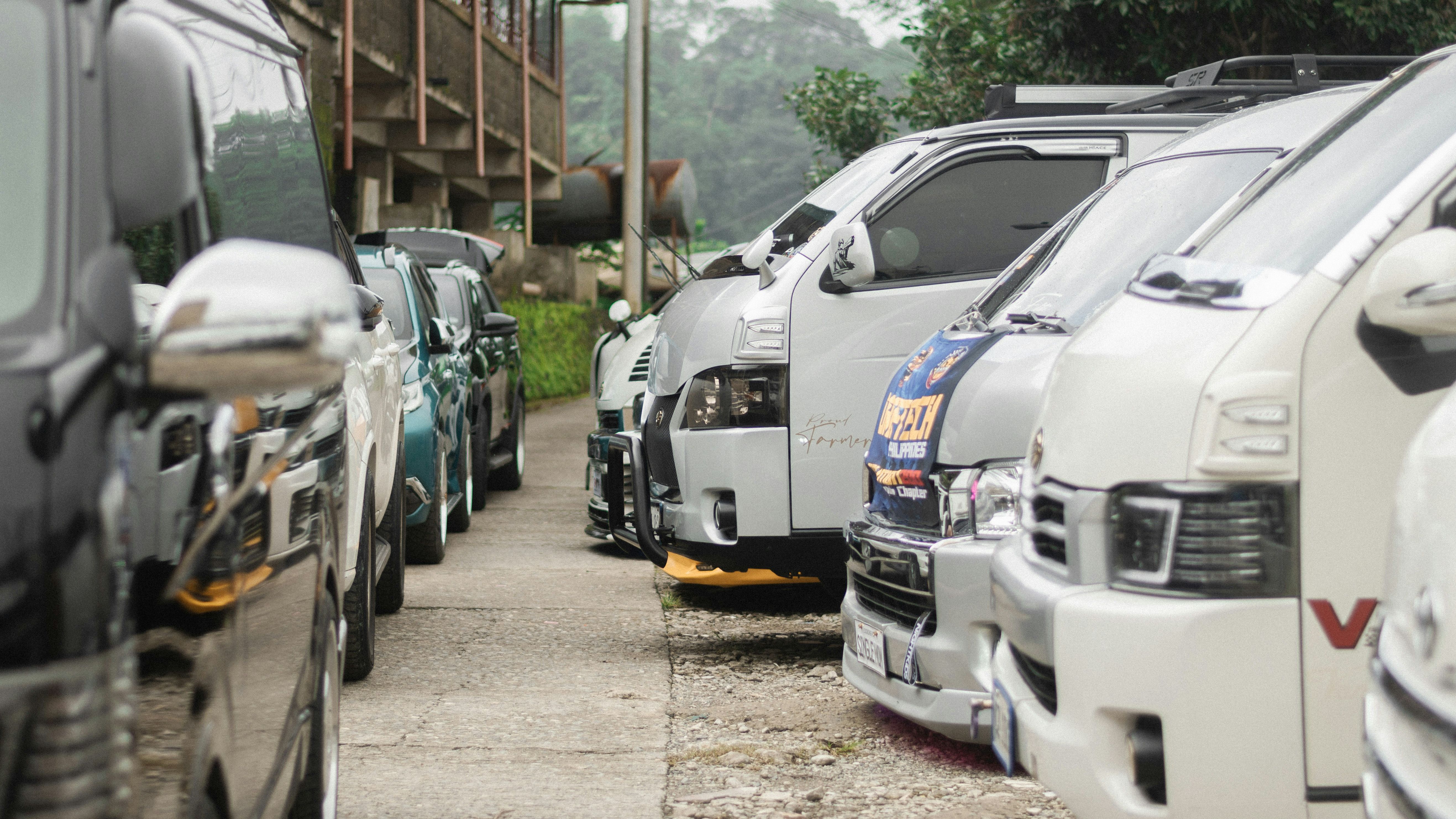 A row of customized vans and vehicles, including Toyota Hiace models, parked closely together in an outdoor setting with greenery in the background.