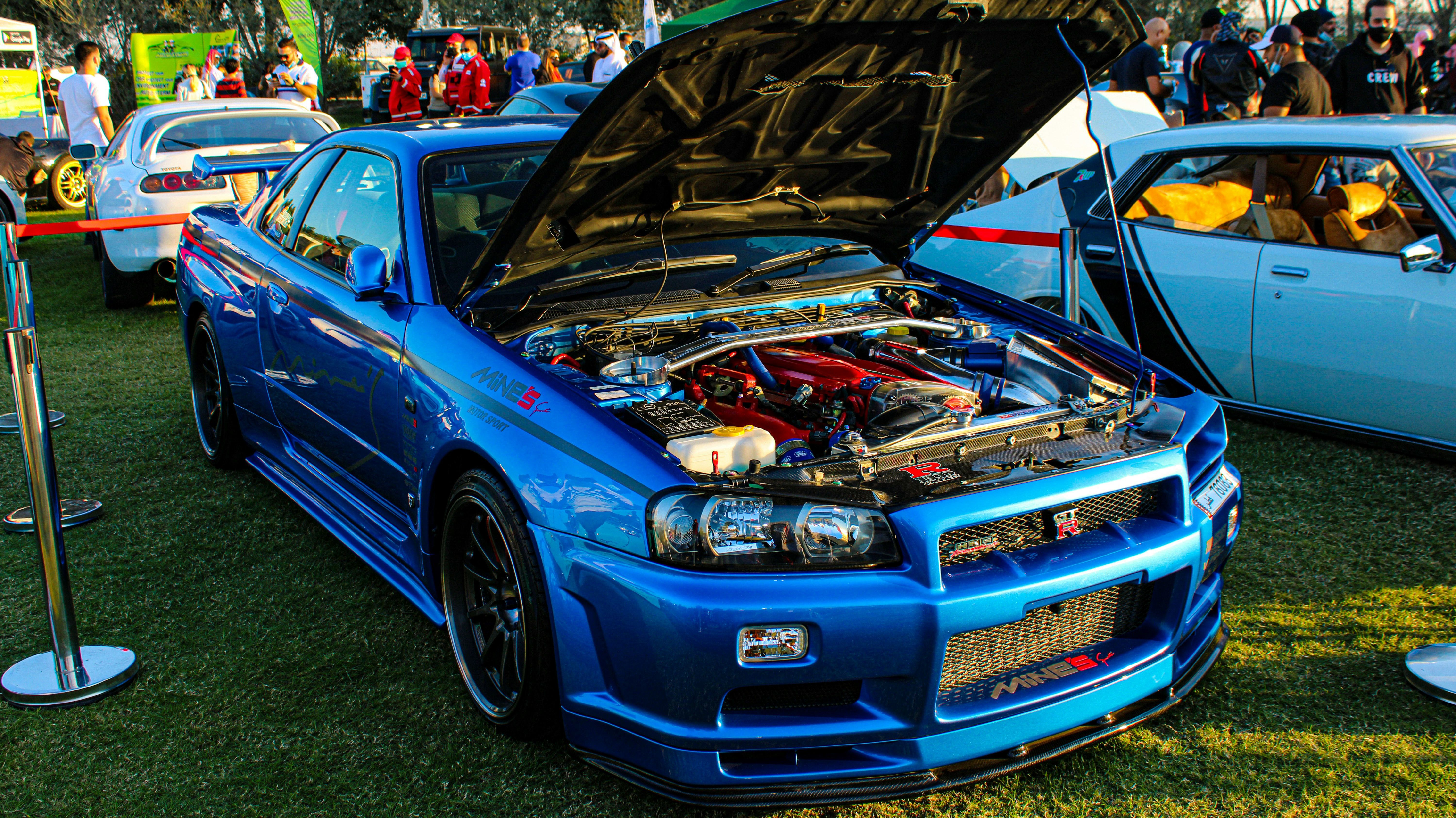 Blue Nissan Skyline GT-R R34 with hood open, showcasing the engine at a JDM car show.