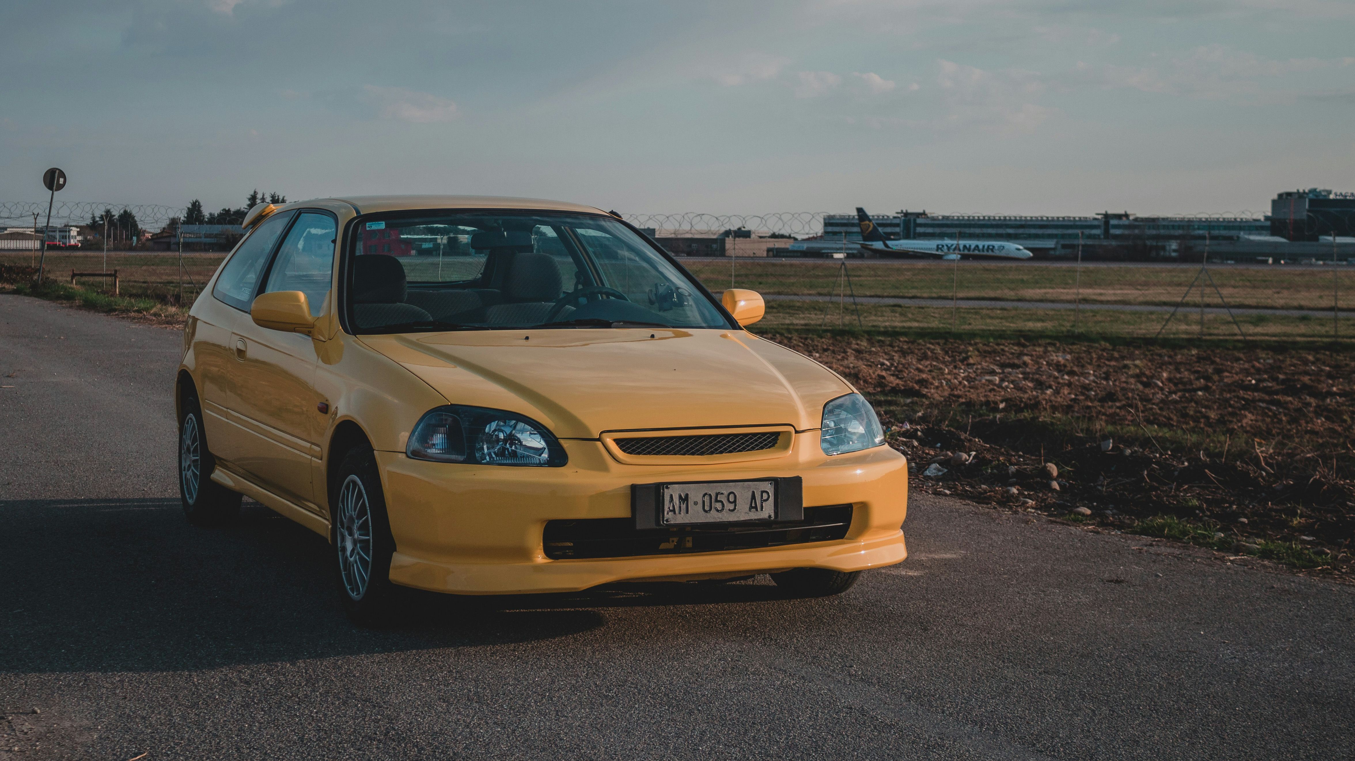 Yellow Honda Civic hatchback parked near an airport, embodying JDM style with a clean front lip and sporty look.